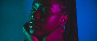 A stylish woman of colour stands against a blue background as brightly colored lights reflect off of her healthy skin
