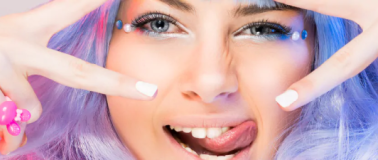 A youthful woman with blue and purple hair sticks out her tongue while flashing the peace sign with her hands.